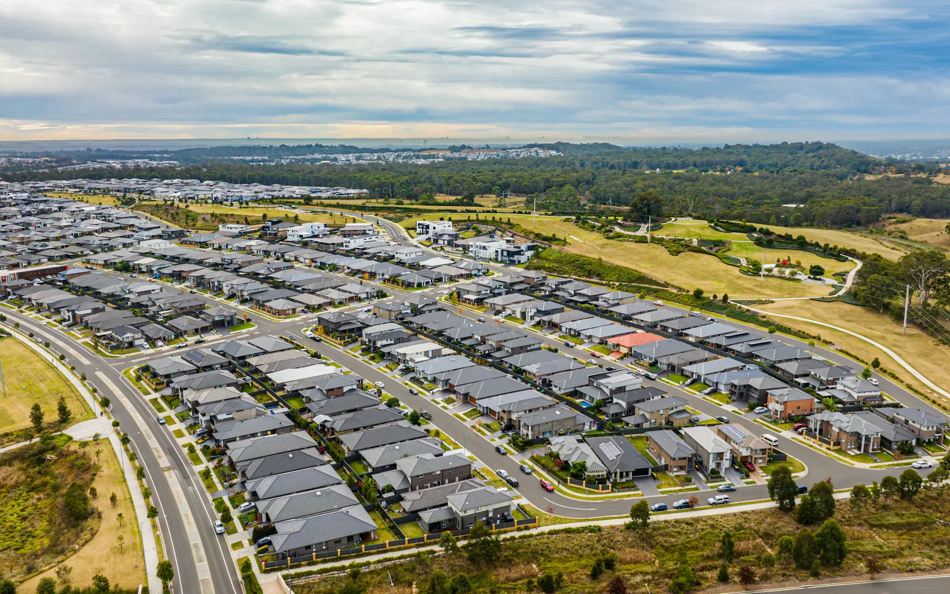 An aerial view of an outer-suburban growth corridor estate: streets of new homes running to the edge of the built-up area, with open paddocks, parkland and bushland still undeveloped beyond it and a distant city skyline on the horizon