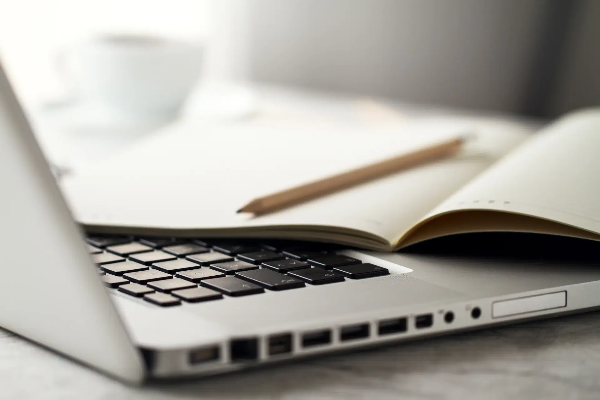 An open laptop on a desk beside a notebook, a pencil and a cup of coffee